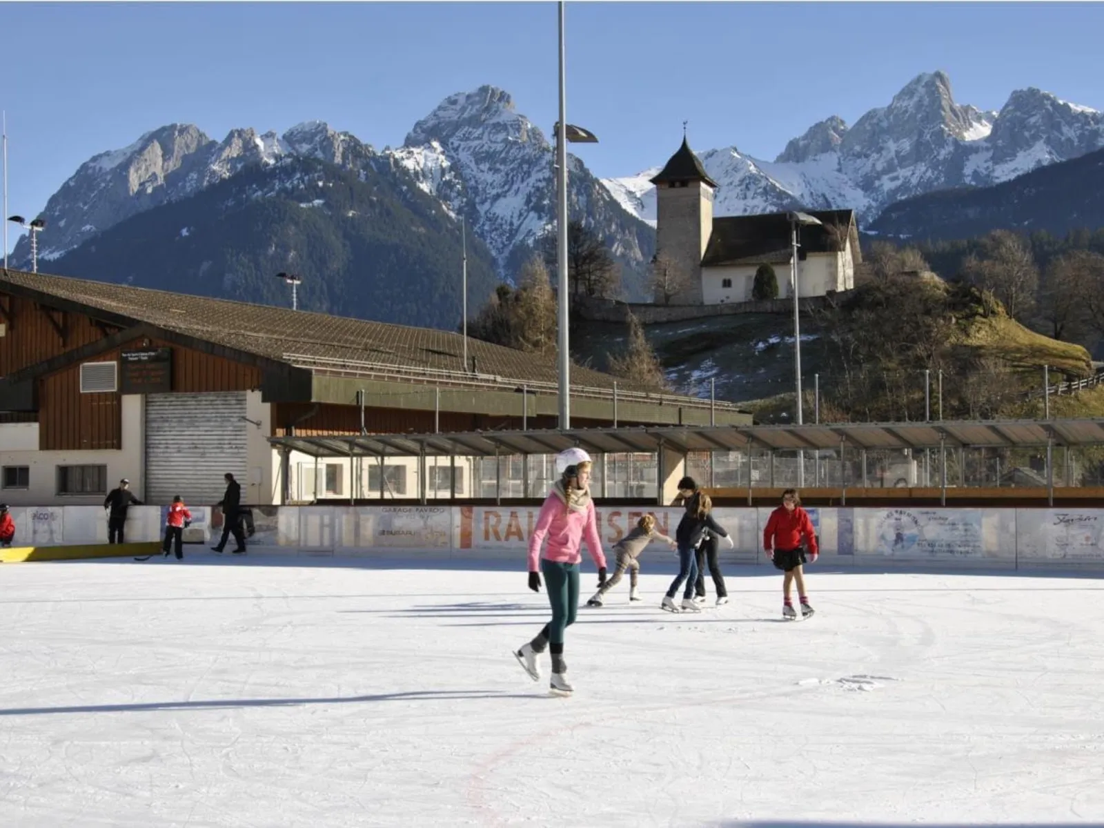 Skaters across the open-air Château-d'Œx rink with a chalet and the old castle tower below snowy peaks. — Patinoire de Château-d'Oex