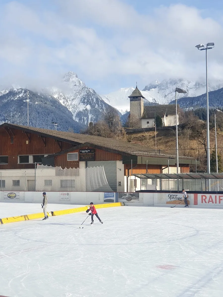 A skater crossing the Château-d'Œx outdoor rink with chalets along the far edge and mountains behind. — Patinoire de Château-d'Oex
