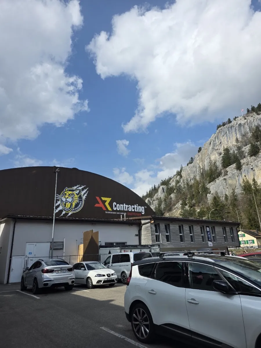 The dark curved-roof Val de Travers ice centre with its logo above the entrance and a limestone cliff behind the car park. — Patinoire de Fleurier (Centre de sports de glace)