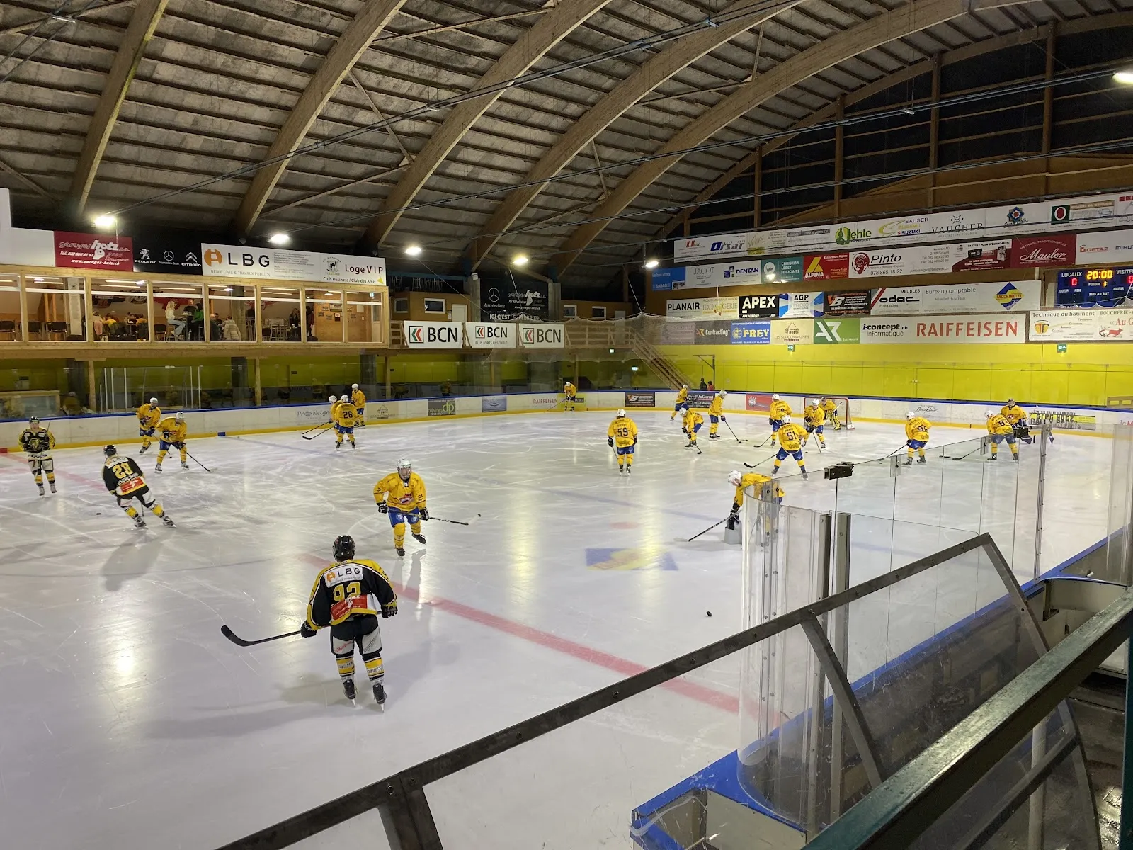 Players spread across the Val de Travers ice under the curved roof with a few skaters near the boards. — Patinoire de Fleurier (Centre de sports de glace)