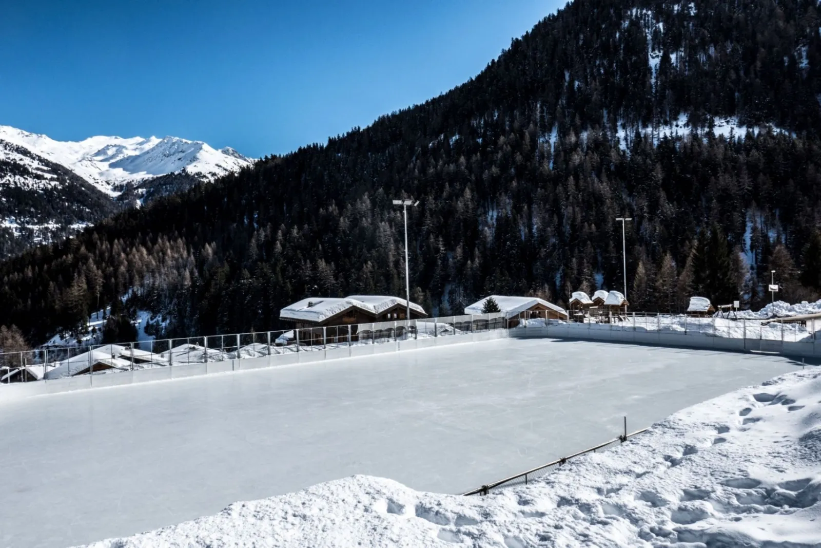 The snow-covered outdoor rink with chalets along its far side and a larch-wooded mountainside behind. — Patinoire de Grimentz