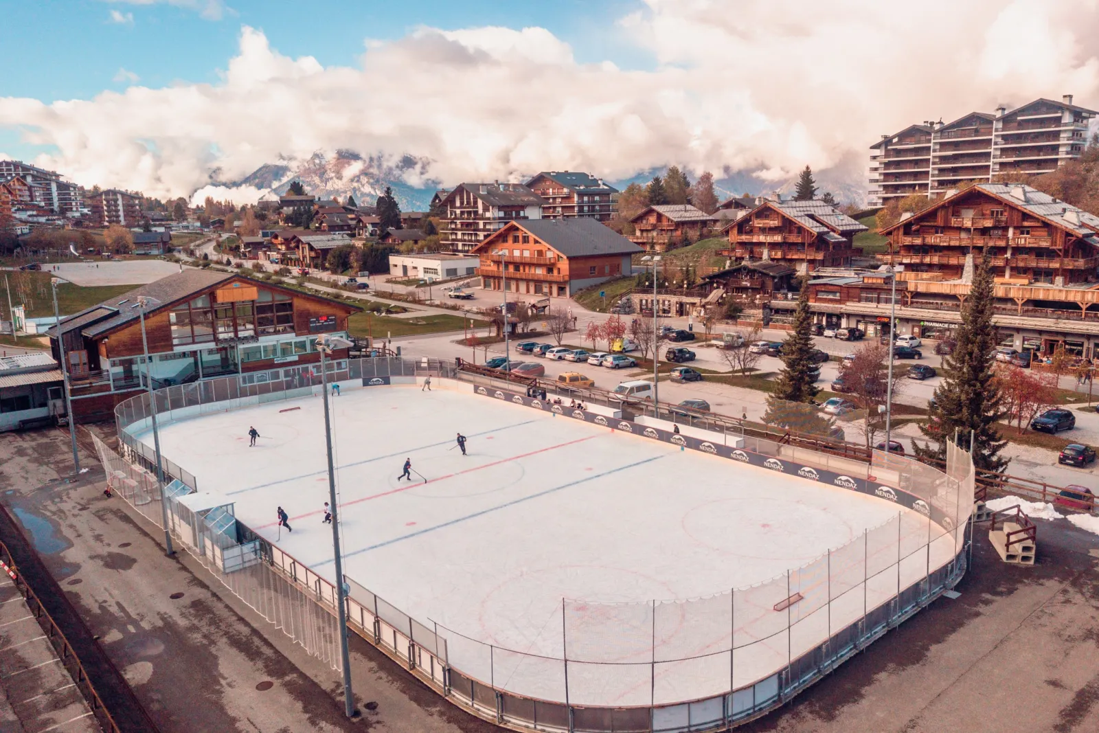 The open-air Haute-Nendaz rink seen from above, a few skaters on the ice and the village chalets and mountains behind. — Patinoire de Haute-Nendaz