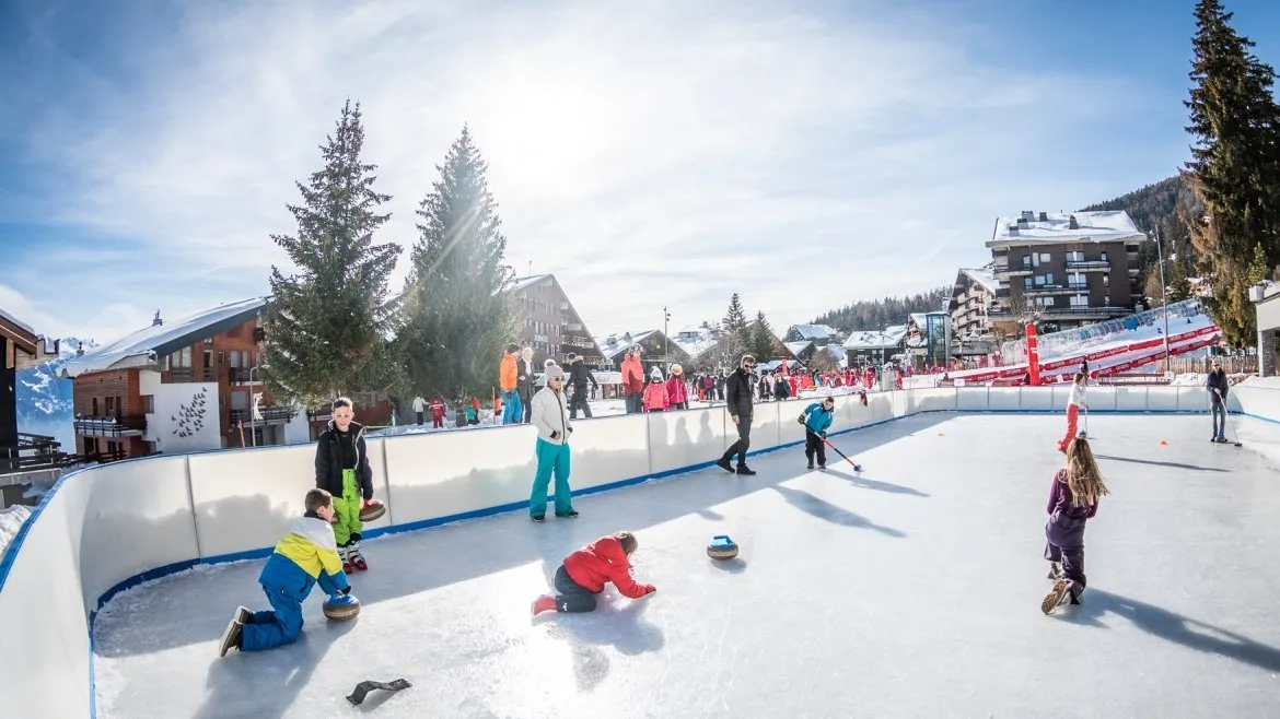 The village-square rink in Anzère in winter, with children playing curling on the ice and chalets and fir trees behind — Patinoire de la Place du Village