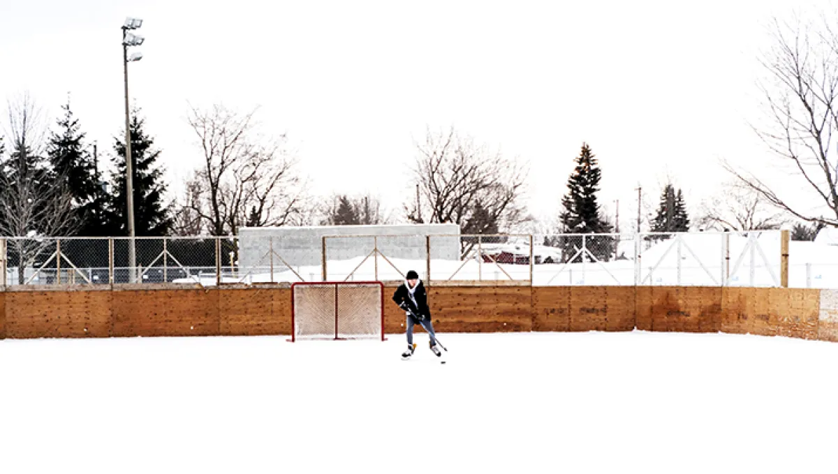 The Patinoire de la Source in Mascouche in winter: a player with a stick in front of a goal at the wooden boards of the outdoor rink, with a floodlight mast and bare trees behind. — Patinoire de la Source (Mascouche)