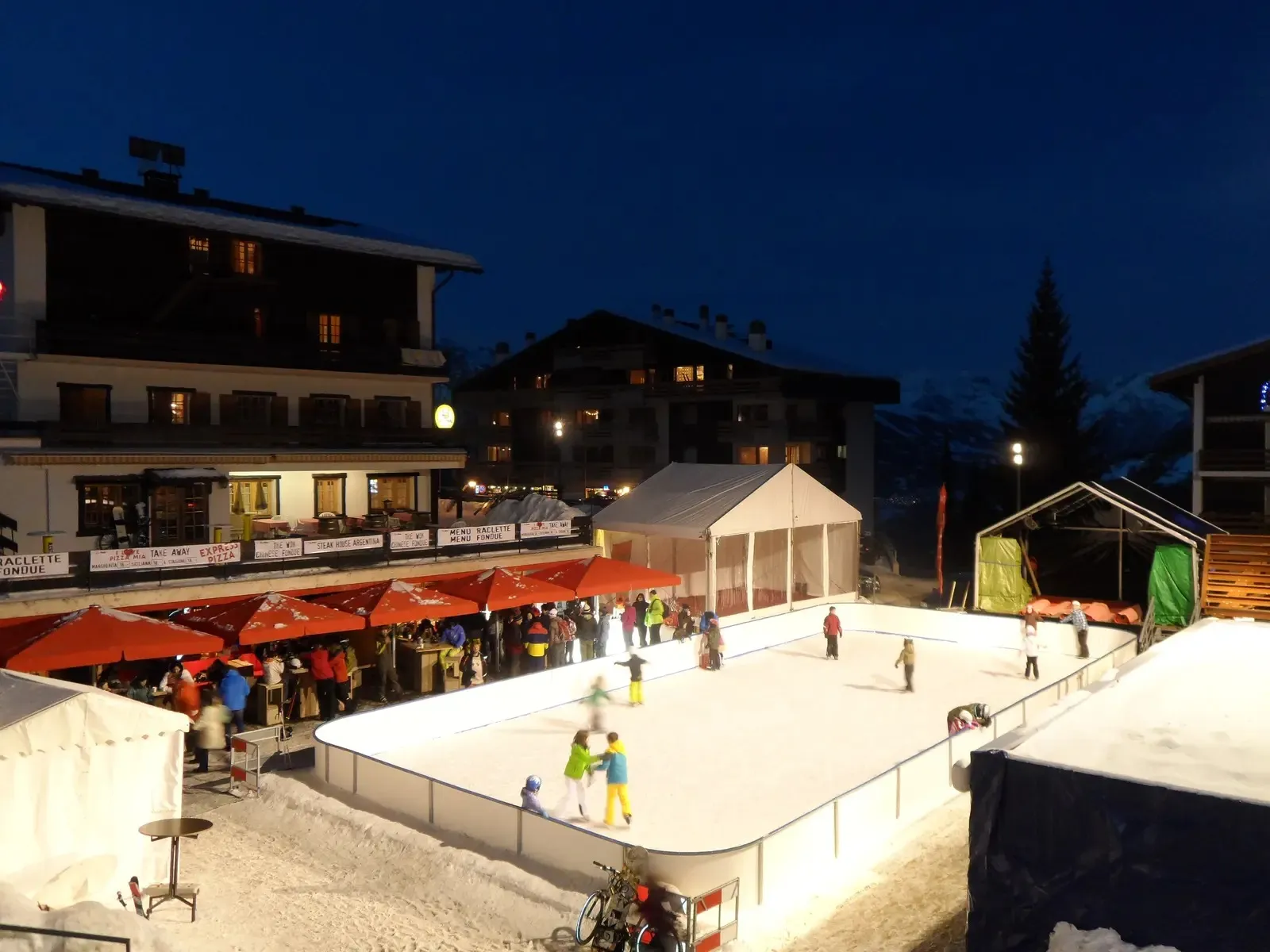 The village rink in La Tzoumaz at dusk, skaters on the lit ice with market stalls, tents and chalets around it. — Patinoire de La Tzoumaz