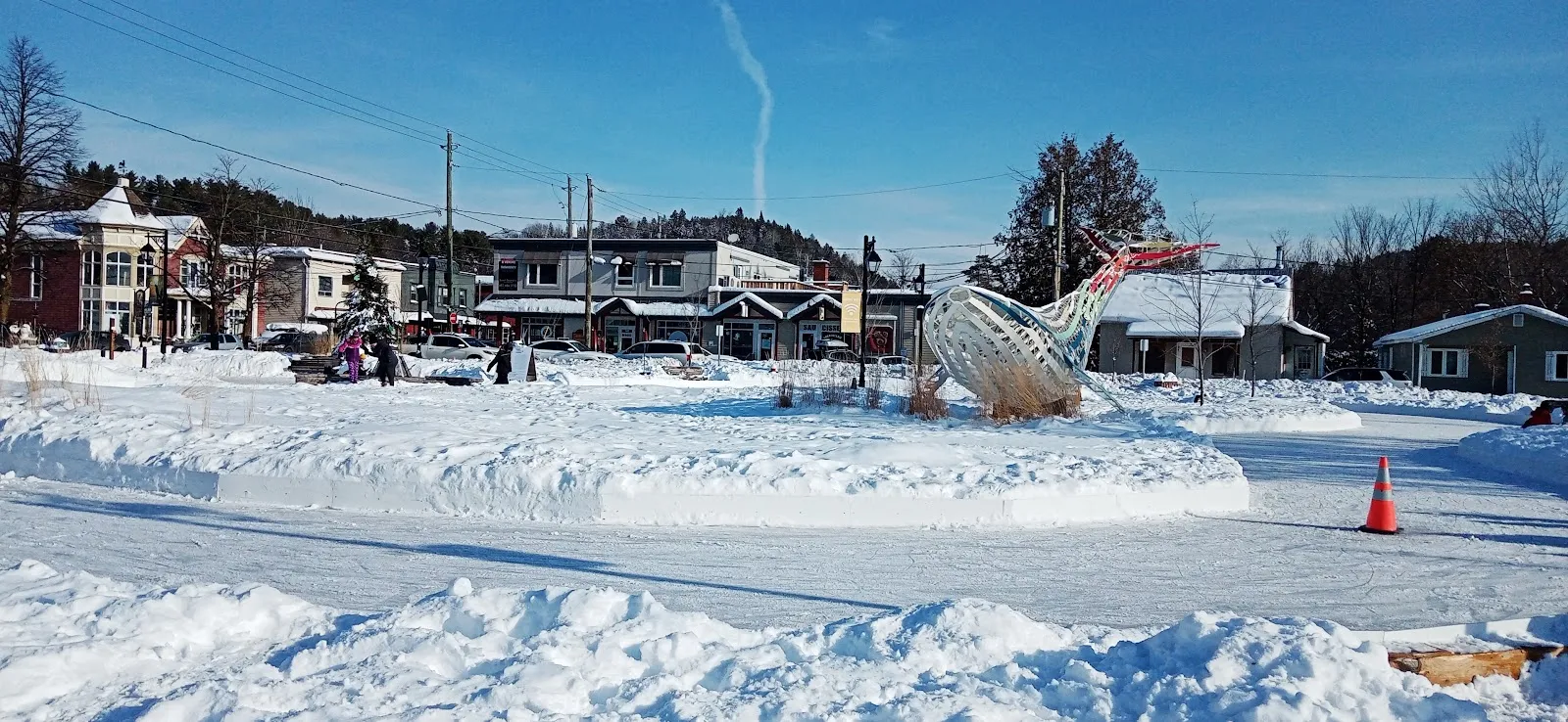 The ice ring of the Espace public in Mont-Tremblant village, banked with snow, with the village buildings and hills behind — Anneau de glace de l'Espace public