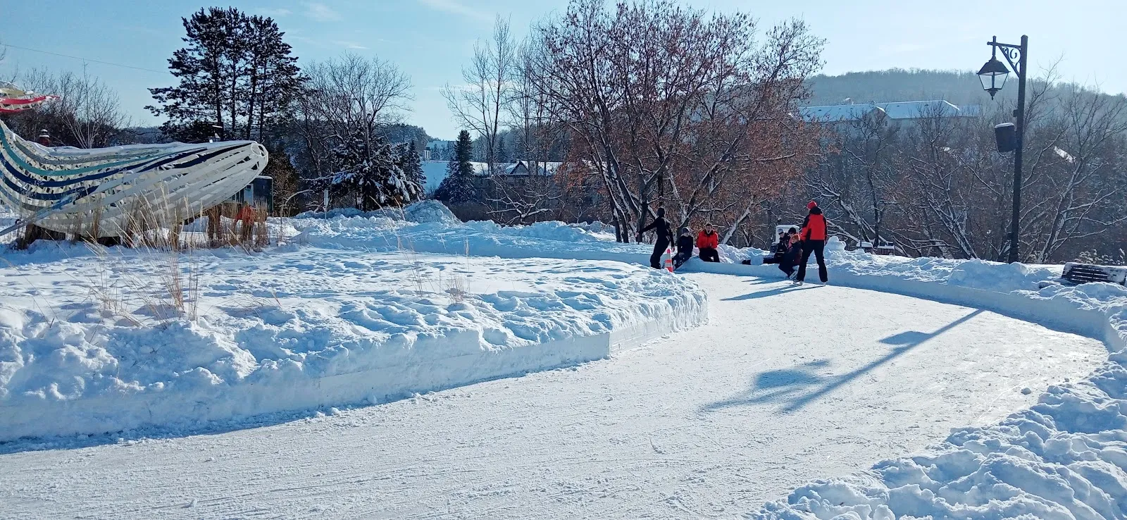 Skaters at a distance on the curving ice ring of the Espace public in Mont-Tremblant village, with a snow-banked edge and sculpted whale — Anneau de glace de l'Espace public