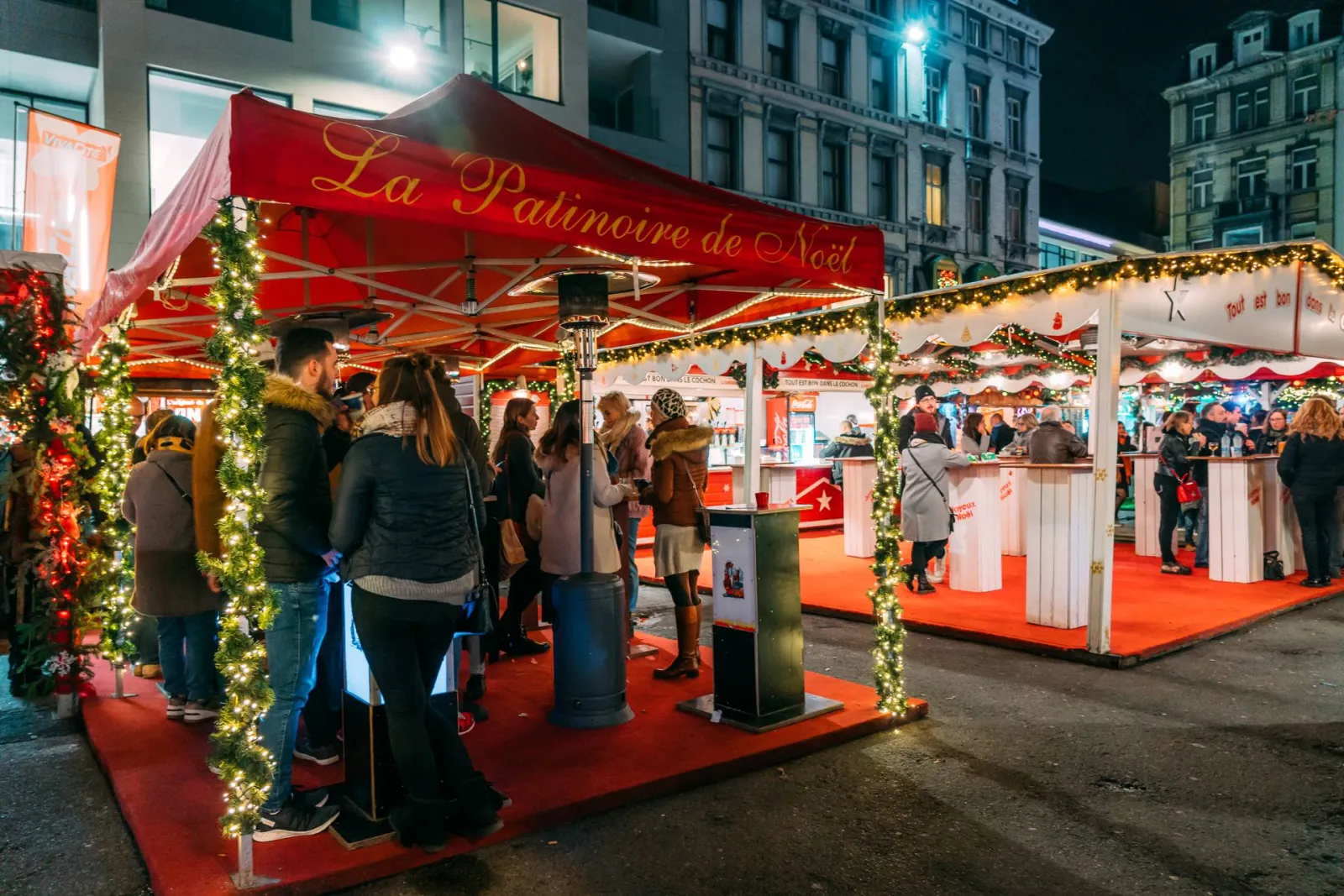 Het rood overkapte paviljoen 'La Patinoire de Noël' voor schaatsverhuur en tickets op de Place de la Cathédrale 's avonds, met wachtende bezoekers; niemand poseert. – Patinoire de Noël de Liège