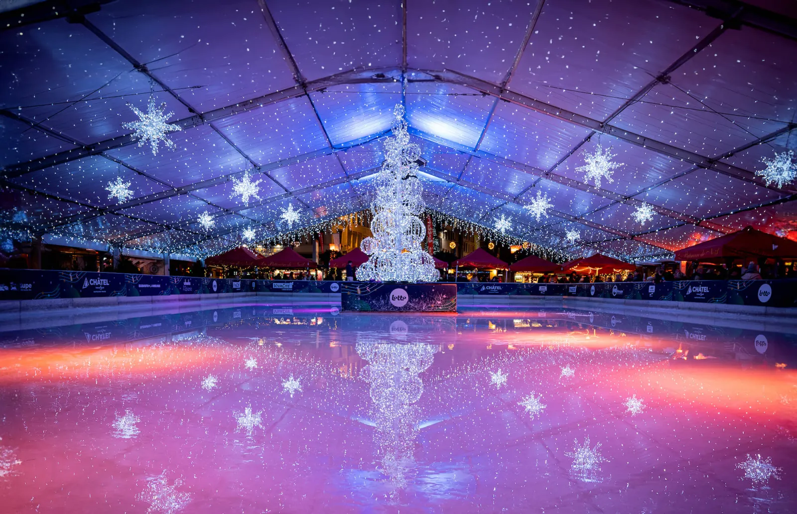De overdekte ijsbaan van de kerstmarkt met haar verlichte plafond, waarin een sneeuwvlok- en kerstboomdecoratie in het ijs weerspiegelt; geen mensen. – Patinoire de Noël de Liège