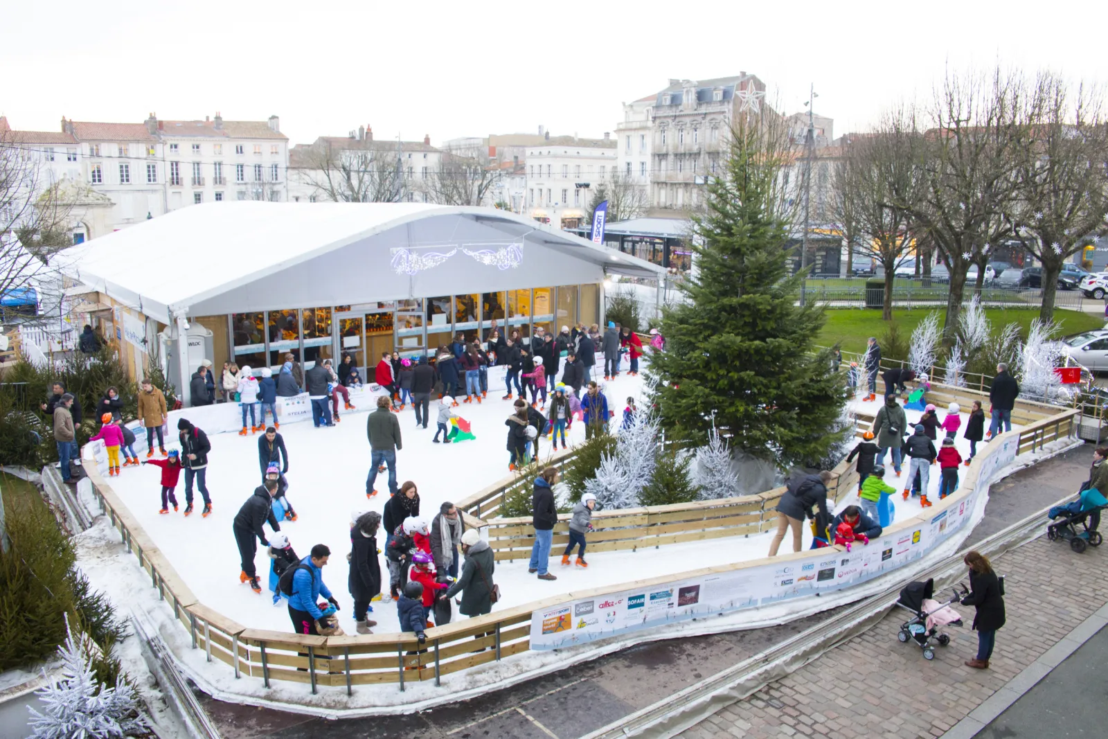 The Rochefort Christmas rink on the place Colbert seen from above, with skaters spread across the white sheet inside wooden boards, fir trees along the edge and a white event tent behind. — Patinoire de Noël de Rochefort (place Colbert)
