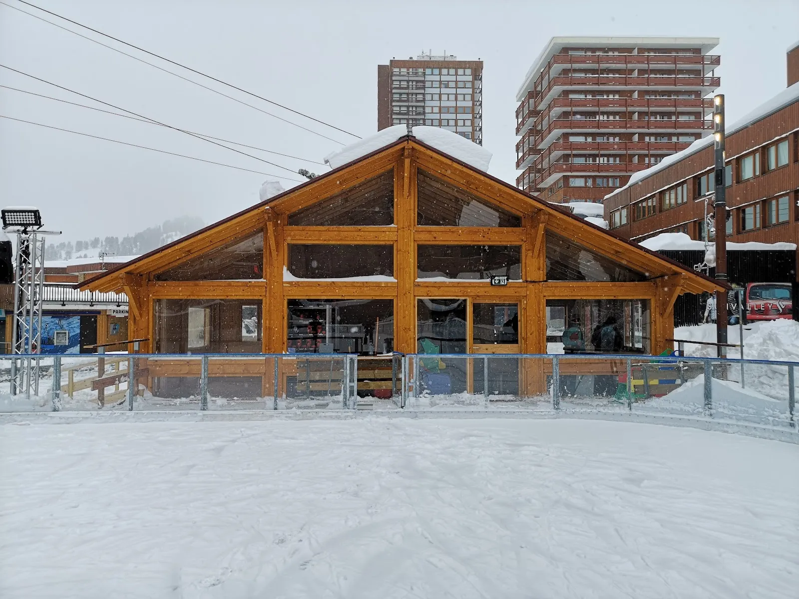 The timber chalet building of the open-air rink at Plagne Centre, with its glazed front and rails along the ice, set among snow-covered resort buildings. — Patinoire de Plagne Centre