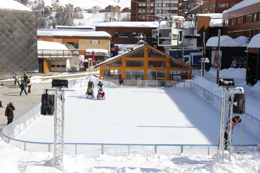 The empty outdoor ice surface of Plagne Centre with its timber chalet behind the boards and the resort buildings above. — Patinoire de Plagne Centre
