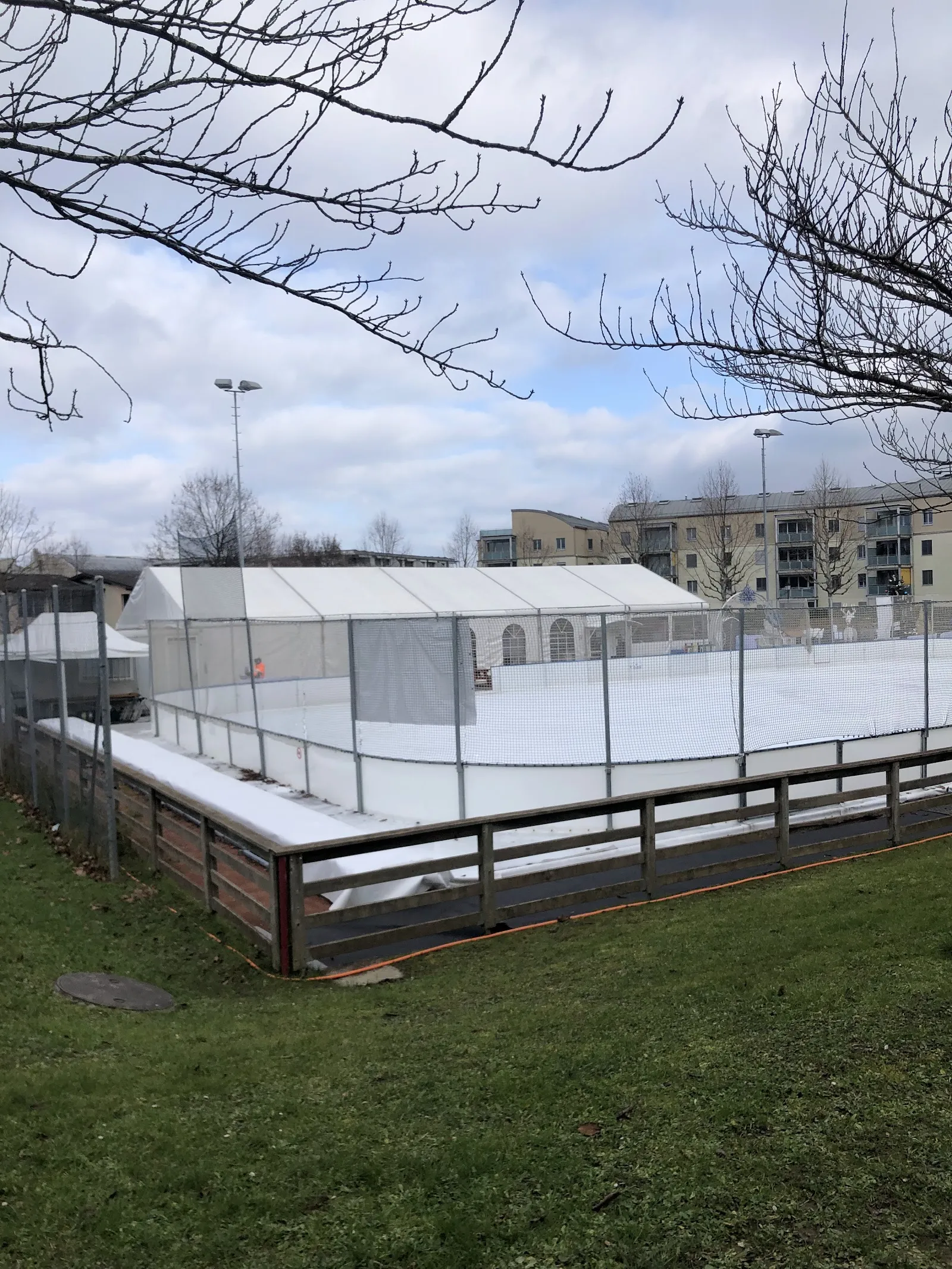 La patinoire d’hiver clôturée de Plan-les-Ouates vue de la pelouse, avec le grand chapiteau blanc à côté de la glace et les immeubles au fond — Patinoire de Plan-les-Ouates