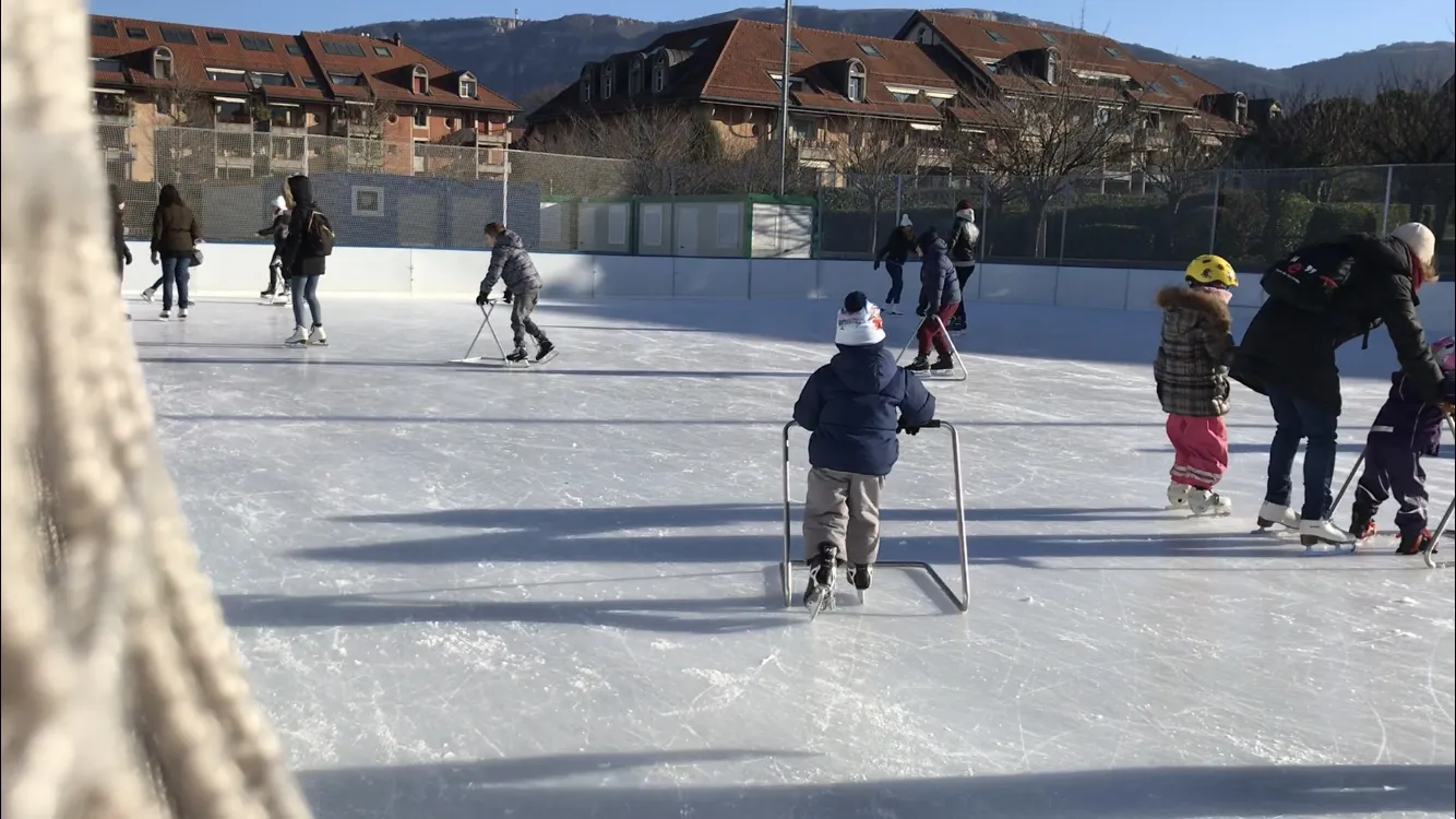Des enfants qui patinent sur la glace extérieure de la patinoire hivernale de Plan-les-Ouates, avec le filet de la piste et les arbres au-delà. — Patinoire de Plan-les-Ouates