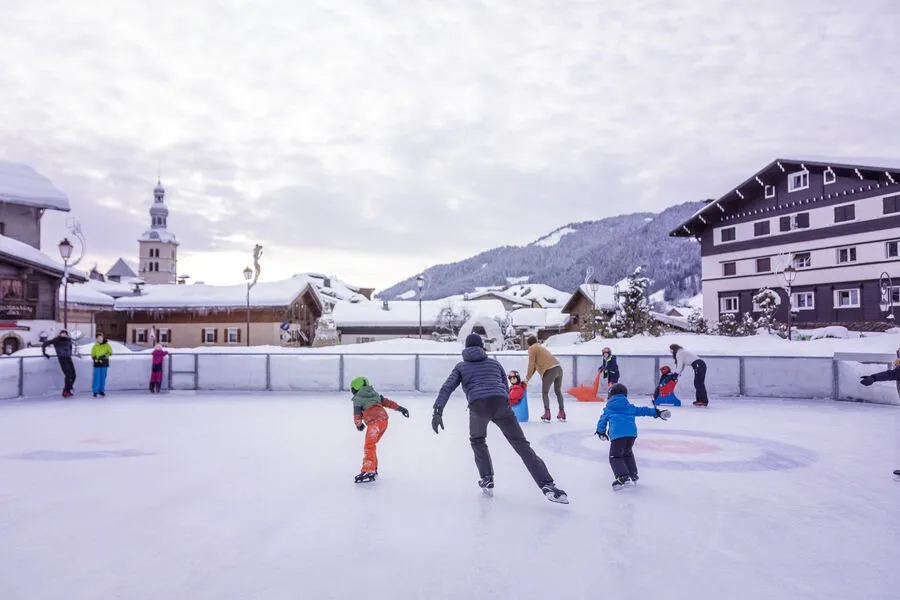 The open-air Megève rink with skaters on the ice, the village's chalets and church tower and the snowy mountains behind. — Patinoire de plein air de Megève