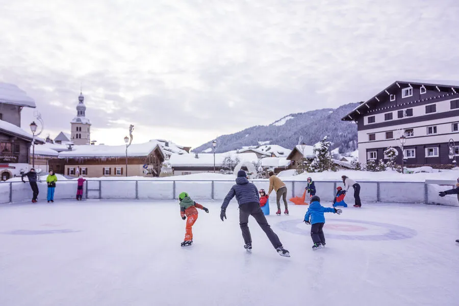 The open-air rink in Megève's village centre with its low boards, skaters on the ice and the church and chalets behind. — Patinoire de plein air de Megève