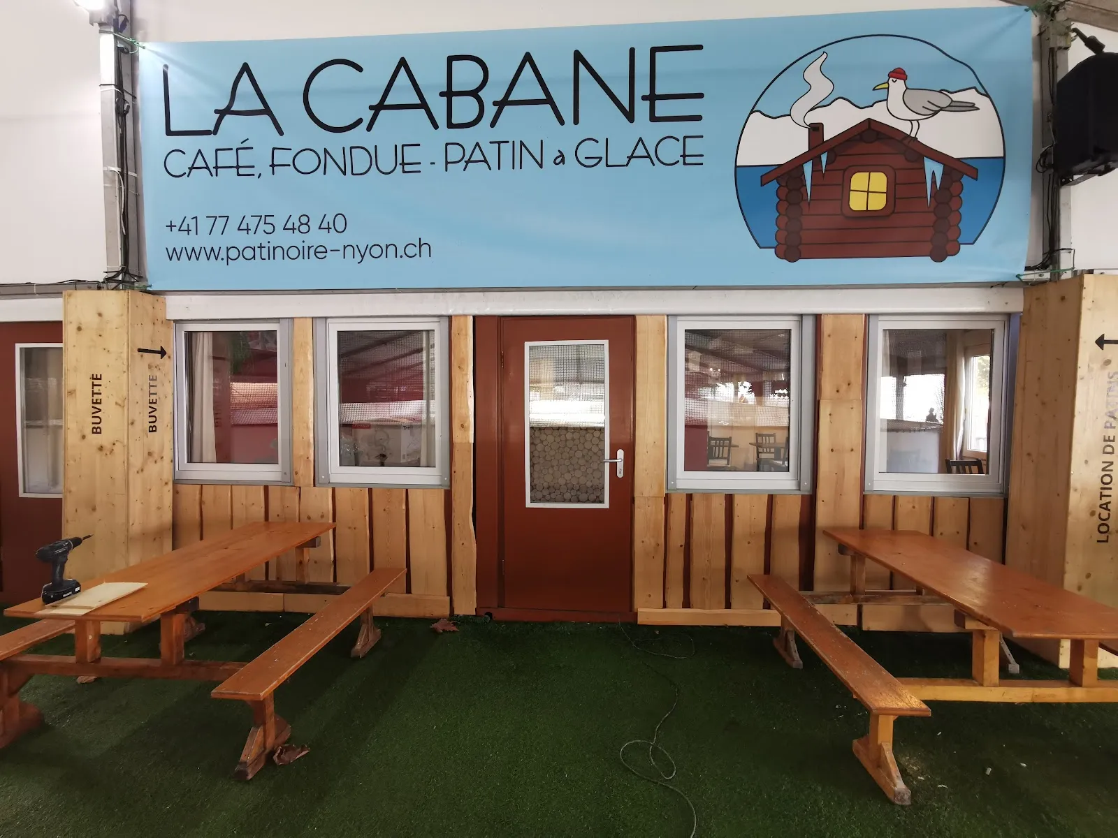 The wooden La Cabane buvette inside the Nyon rink tent, with its banner naming the cafe, fondue and skating, and picnic tables on artificial turf in front — Patinoire de Rive (La Cabane)