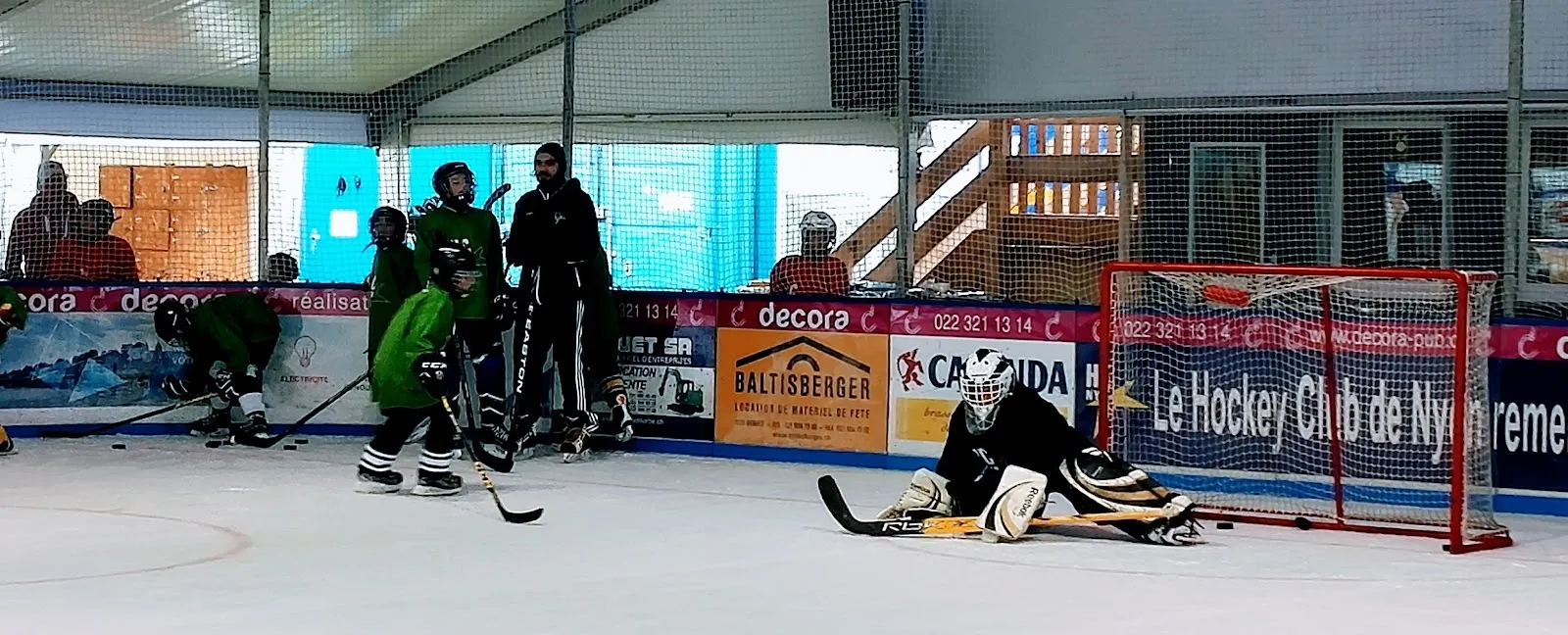 Hockey players at a face-off on the ice of the Patinoire de Rive in Nyon, with the boards and the club's sponsor panels behind. — Patinoire de Rive (La Cabane)