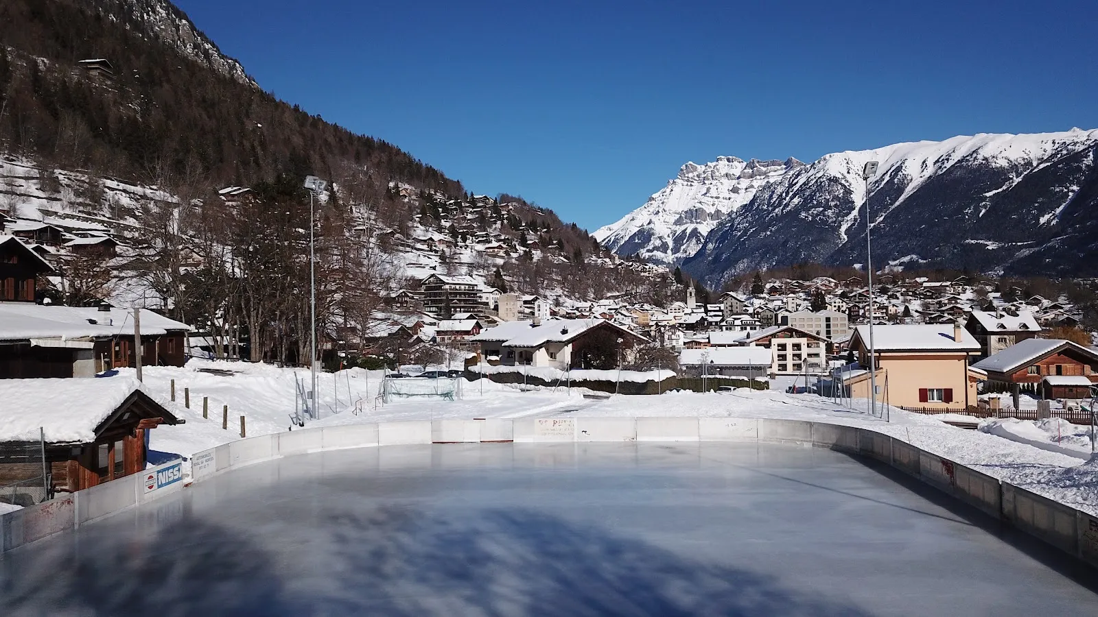 The open-air ice sheet inside its white boards, seen from the end with chalets and snow-covered mountains behind. — Patinoire de Salvan