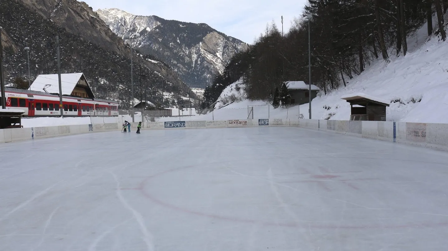 The open-air Sembrancher rink under a light snow cover, with chalets and a forested mountain behind the boards. — Patinoire de Sembrancher