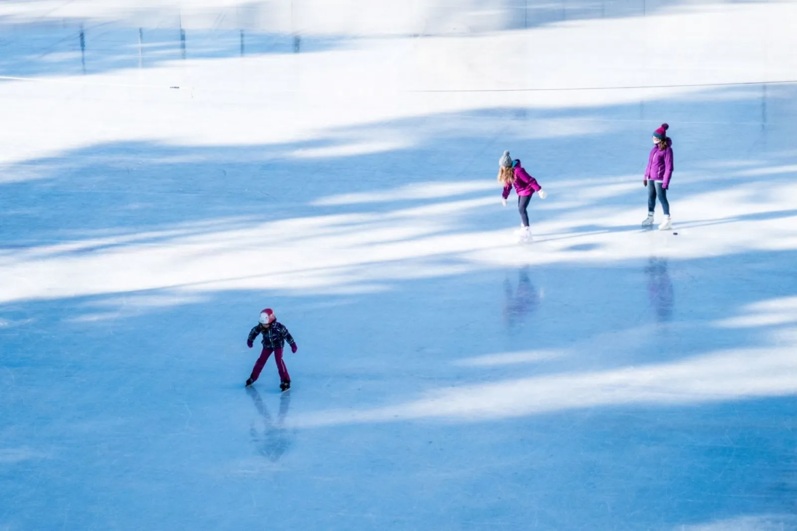 Three skaters on blue-tinted ice in winter sunlight, seen from above. — Patinoire de St-Luc