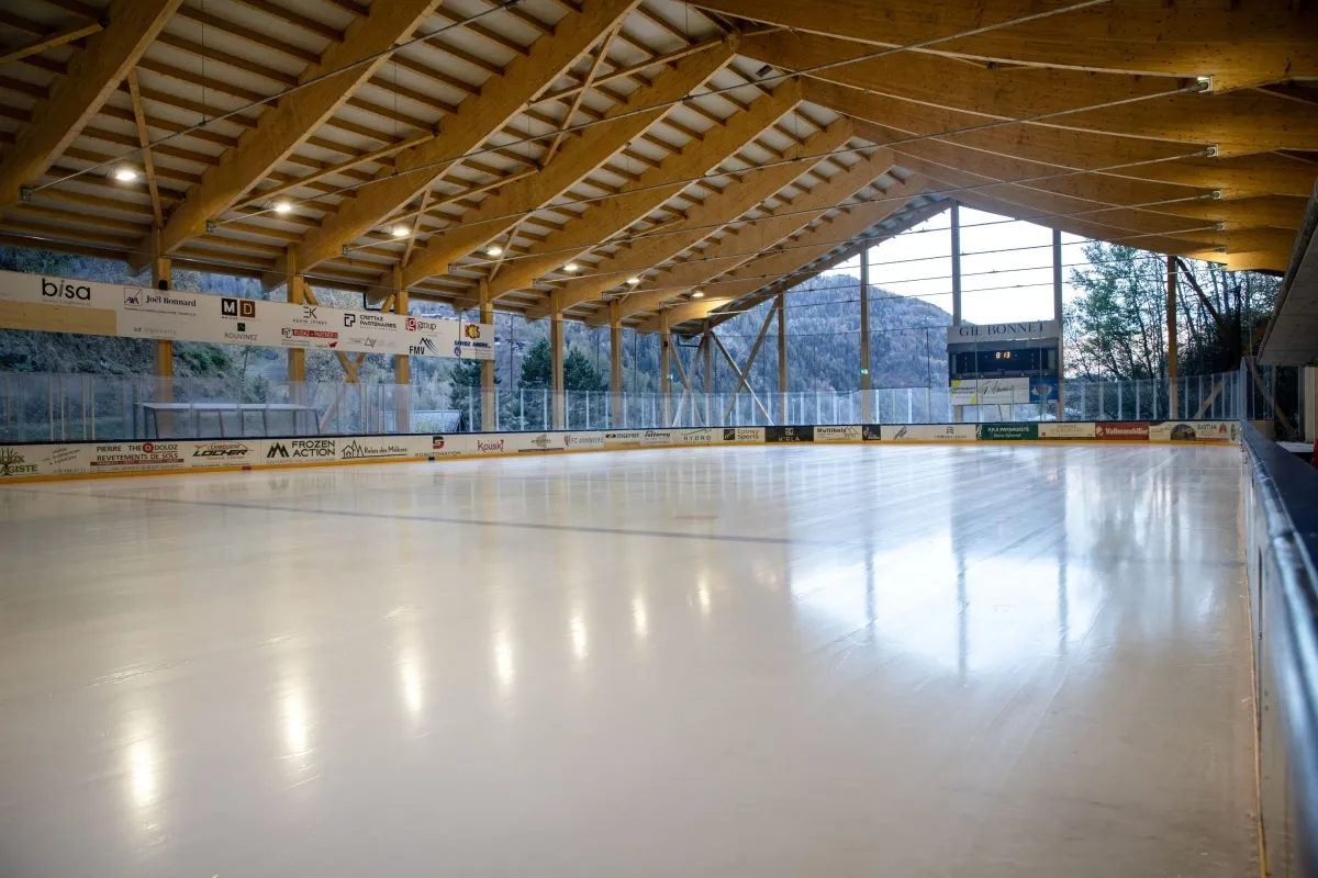 The empty ice sheet under a timber roof, with advertising boards along the side and the wooded valley slope visible beyond the open side. — Patinoire de Vissoie