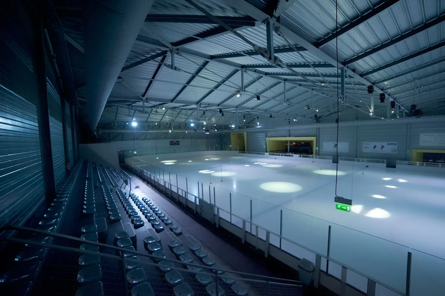 The empty rink seen from the terraces, with the sloping steel roof, the grandstand on the left and spotlights on the ice — Patinoire des 3 Seine