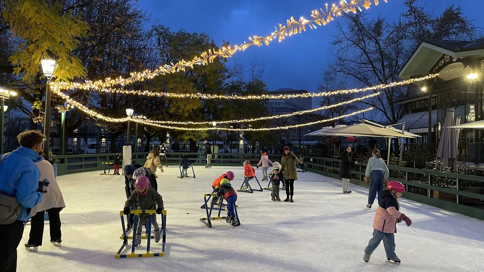 La patinoire en plein air au crépuscule sous des guirlandes lumineuses, toute la surface de glace avec quelques patineurs à moyenne distance et la terrasse au-delà. — Patinoire des Bastions