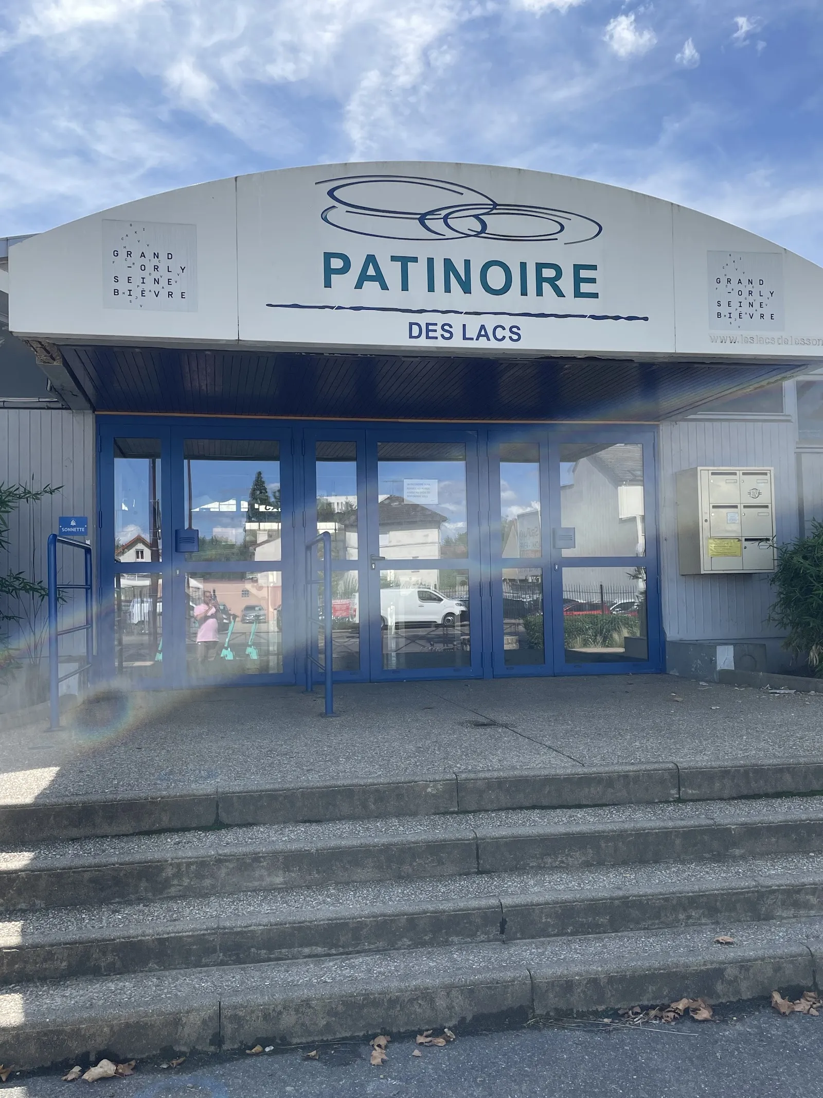 The arched entrance of the Patinoire des Lacs in Viry-Châtillon with its name and ring logo above the blue glass doors and the concrete steps in front — Patinoire des Lacs