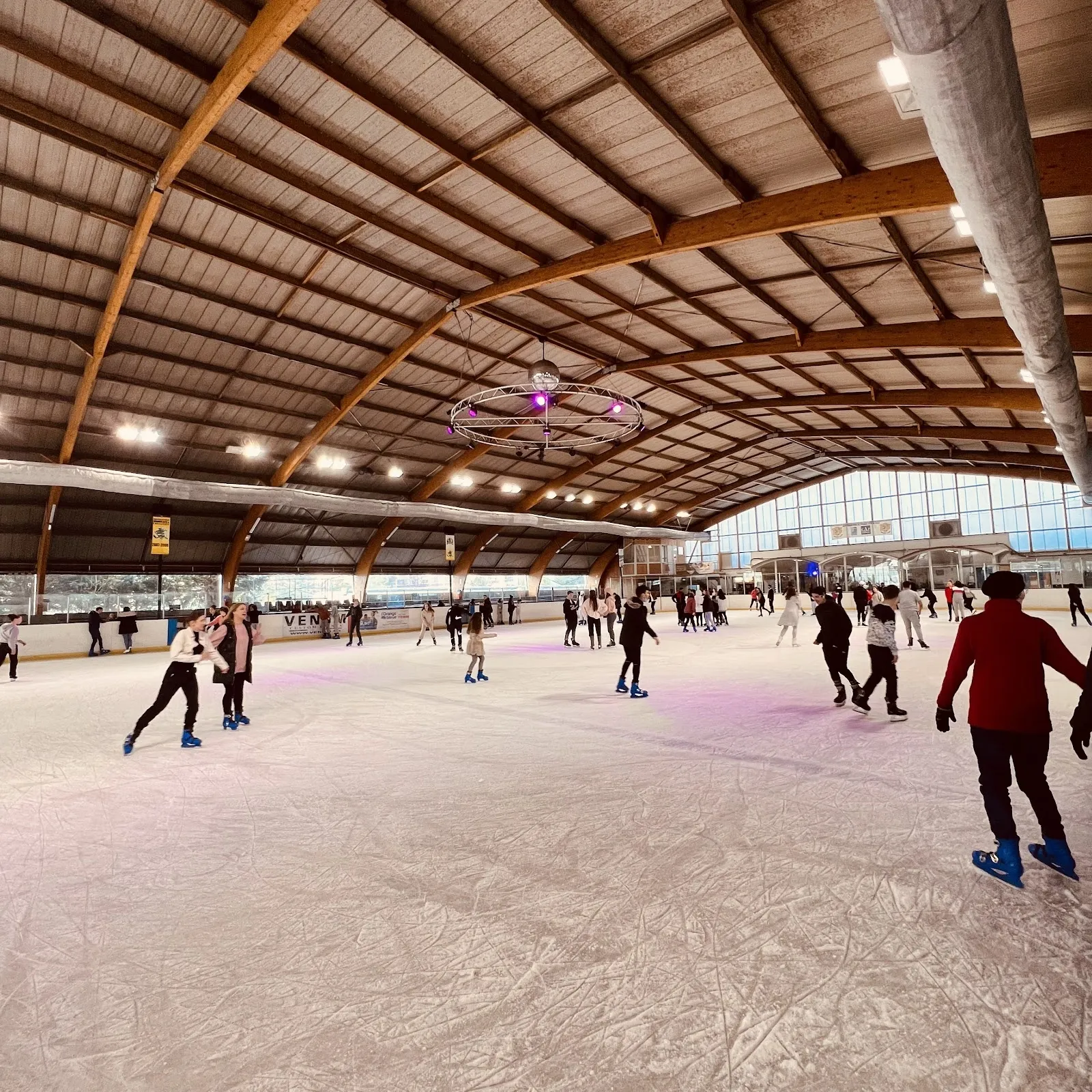 Skaters on the ice sheet in the Viry-Châtillon rink, under the curved timber roof of the hall. — Patinoire des Lacs