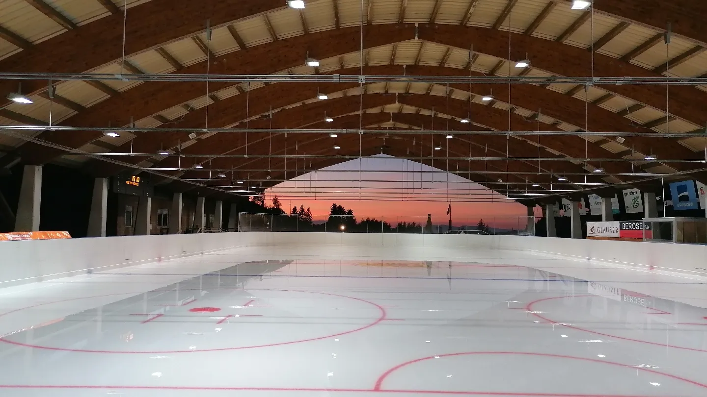 The empty ice under the arched timber roof of the Paccots rink, with the red centre lines and mountain light beyond the open sides. — Patinoire des Paccots