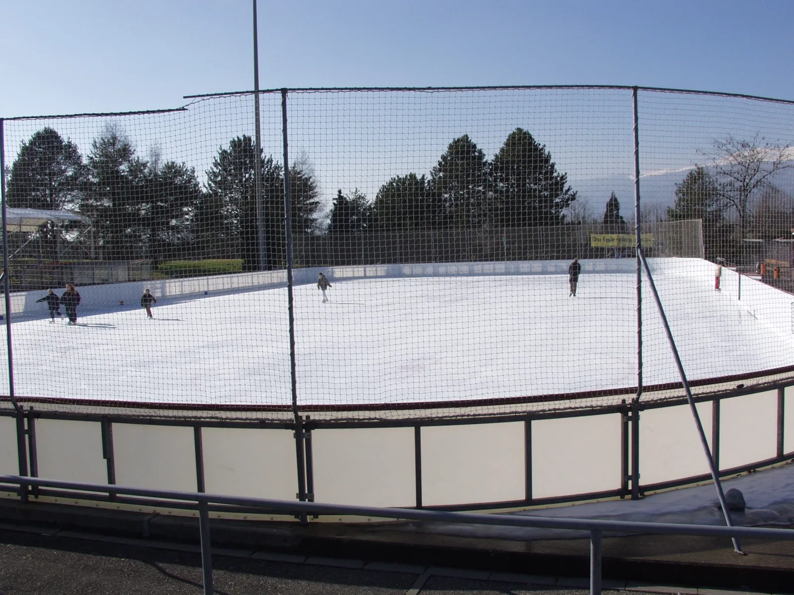 La glace extérieure des Vergers à Meyrin derrière ses bandes blanches et ses filets de sécurité, quelques patineurs au loin. — Patinoire des Vergers
