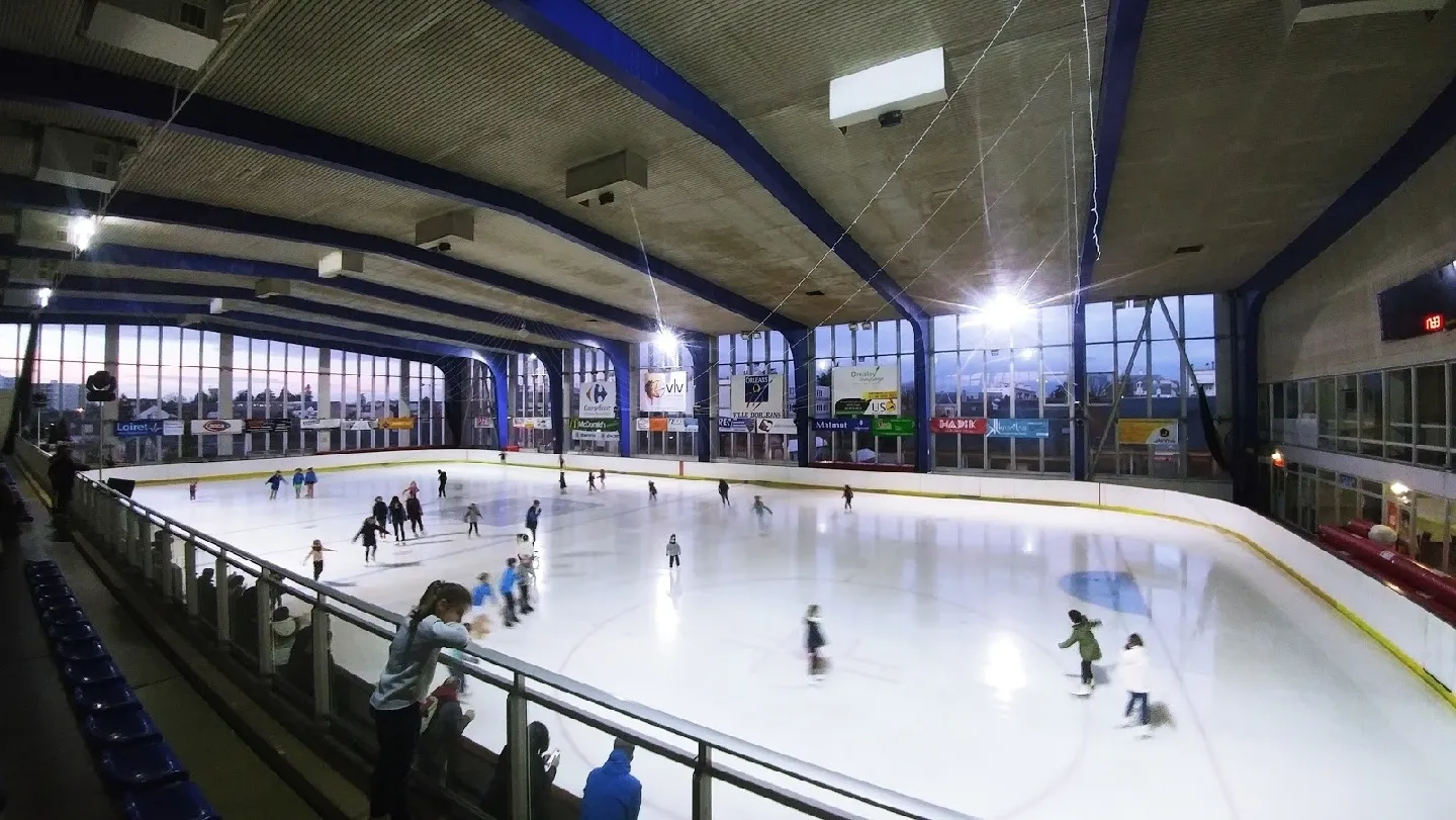 Une séance de patinage public sur la glace de la patinoire d’Orléans, vue depuis les gradins sous la charpente métallique bleue. — Patinoire d'Orléans