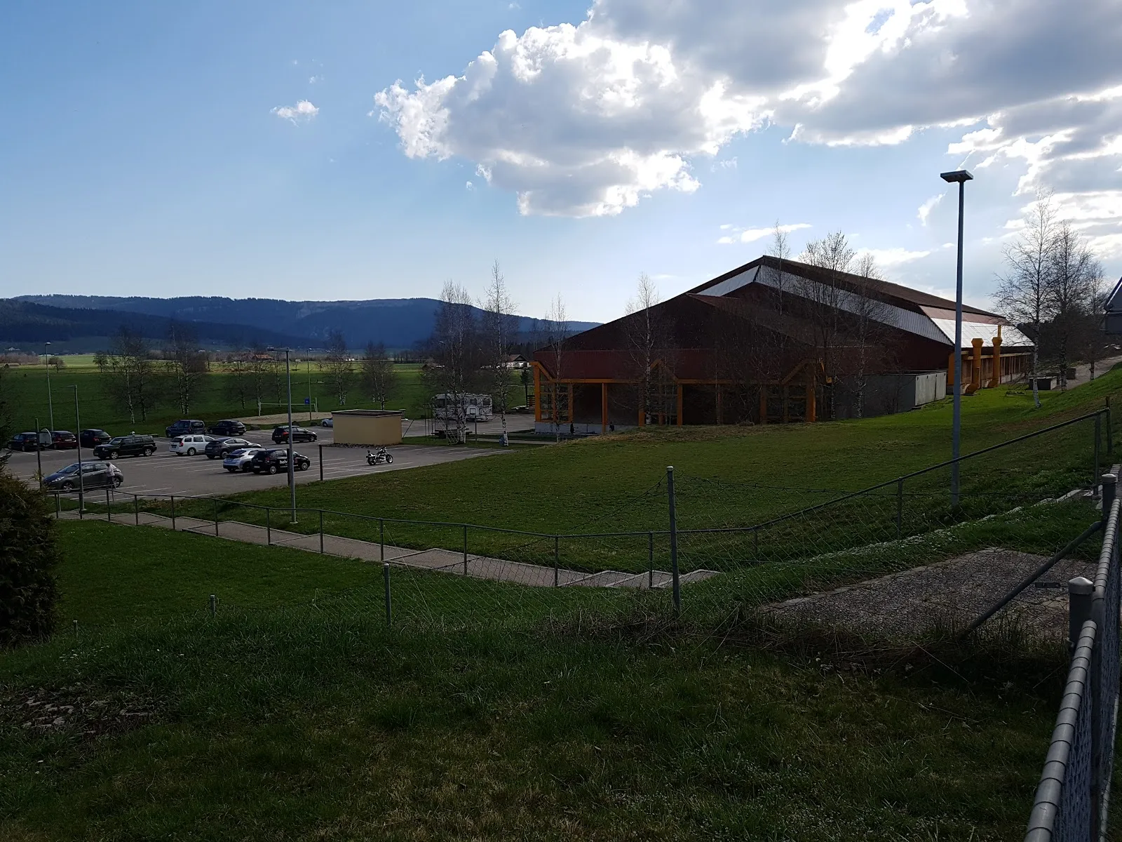 La patinoire du Bugnon, au toit de bois sombre, vue de l’autre côté du pré et du parking, avec les collines du Jura derrière — Patinoire du Bugnon