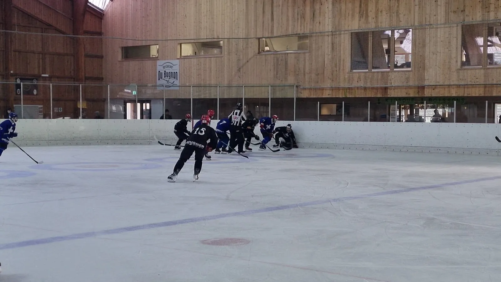 Un joueur qui patine sur la glace de la patinoire du Bugnon aux Ponts-de-Martel, avec la halle en bois et sa charpente derrière les bandes. — Patinoire du Bugnon