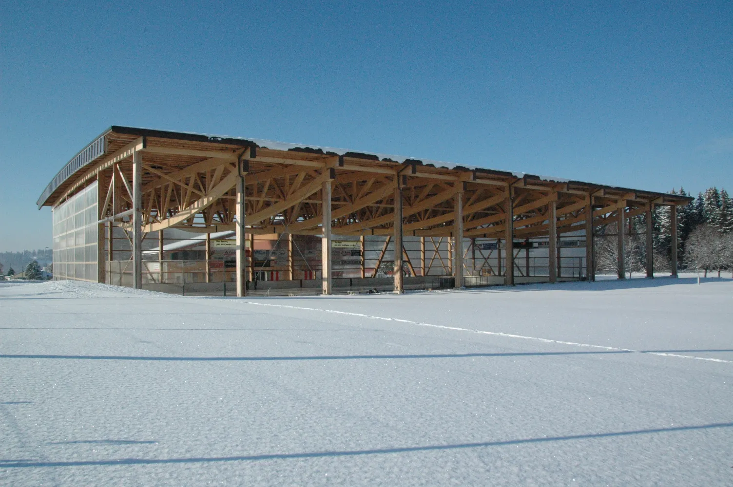 The Communal rink building in snow: a long curved wooden roof on timber posts, open at the sides, with a glazed end wall and a flat snowy field in front. — Patinoire du Communal