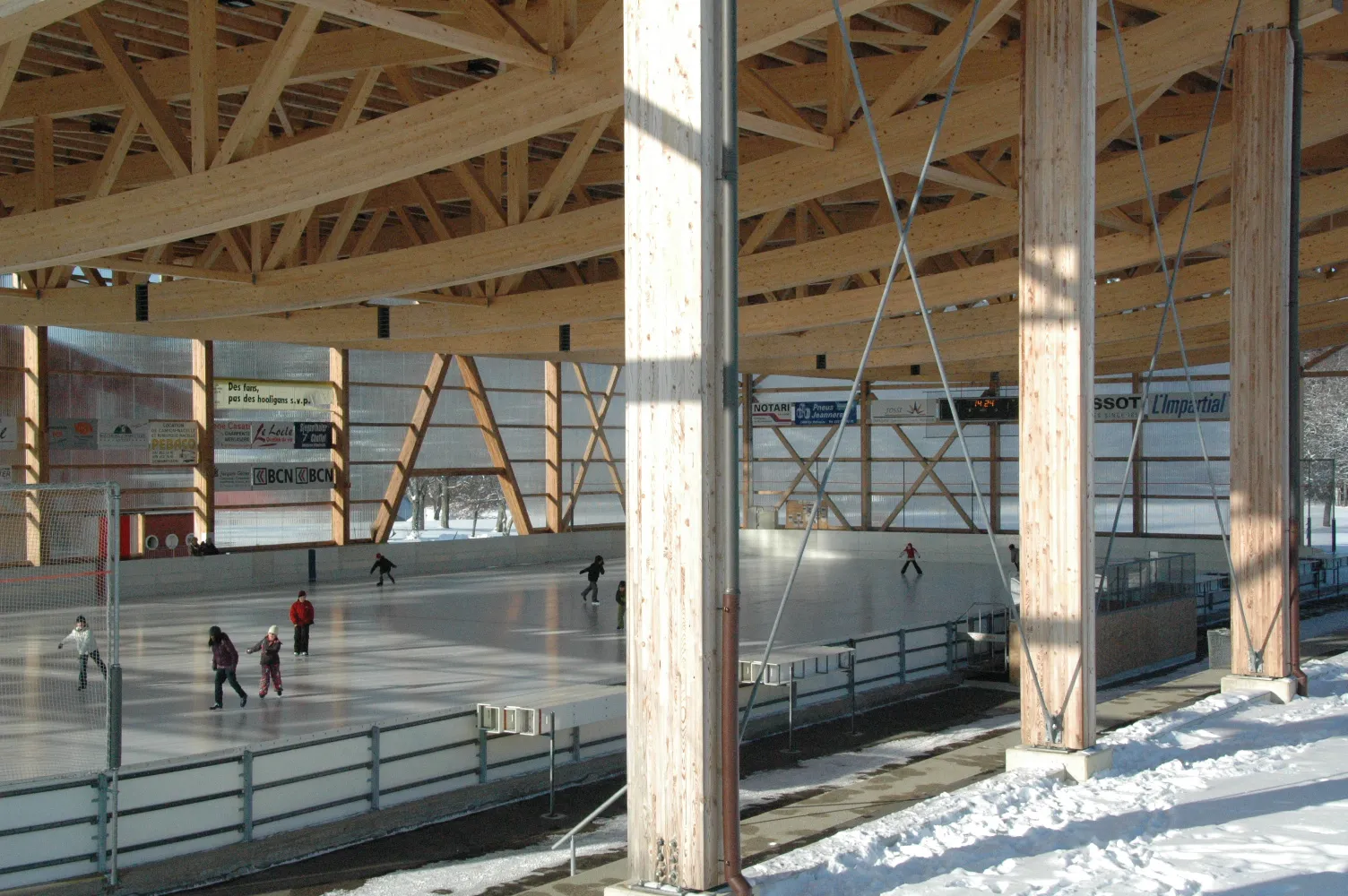Skaters on the ice of the Communal rink under its timber roof beams, seen from the side of the rink in winter light, with the open sides looking onto snow. — Patinoire du Communal