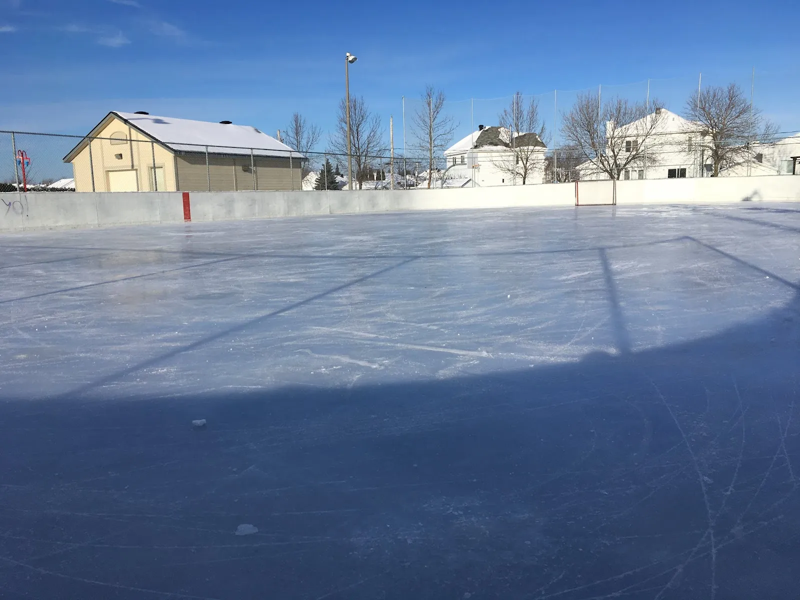 The empty boarded rink at Parc Cardinal in Mirabel on a sunny winter day, with the park chalet and houses behind the boards — Patinoire du parc Cardinal