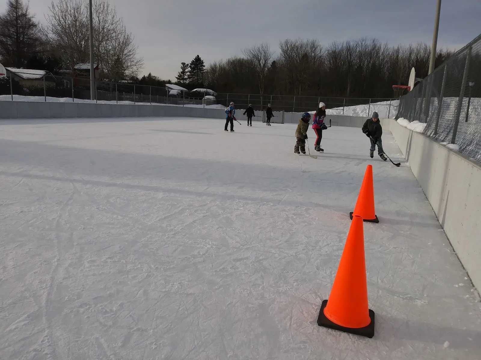 A few skaters at a distance on the ice of the boarded rink at Parc Charlebel in Mirabel, with orange cones and the fence beyond — Patinoire du parc Charlebel