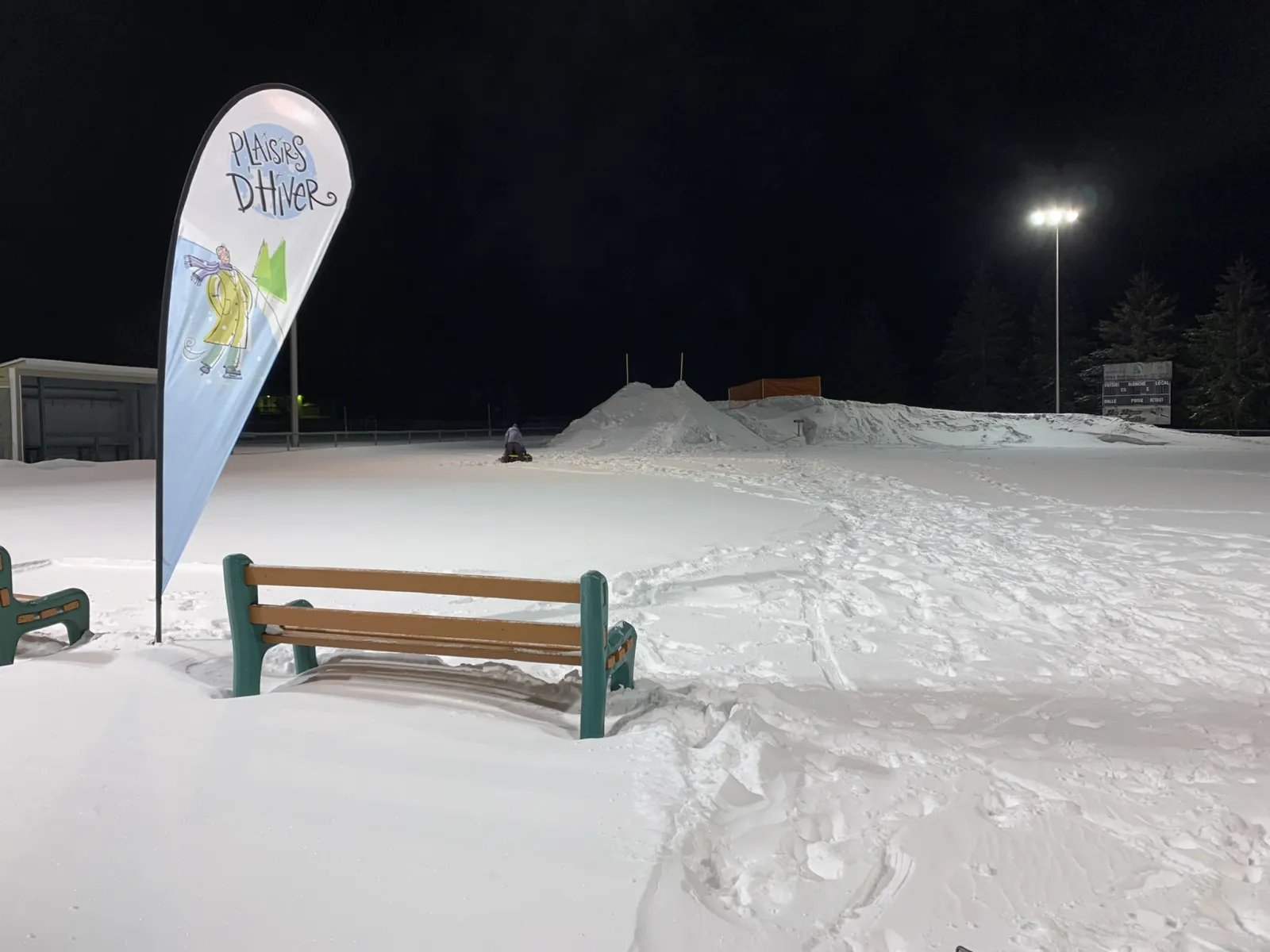 La zona nevada de la pista de noche bajo los focos, con un banco del parque y una bandera de la Maison d'hiver al lado. — Patinoire du parc Claude-Archambault (Saint-Didace)