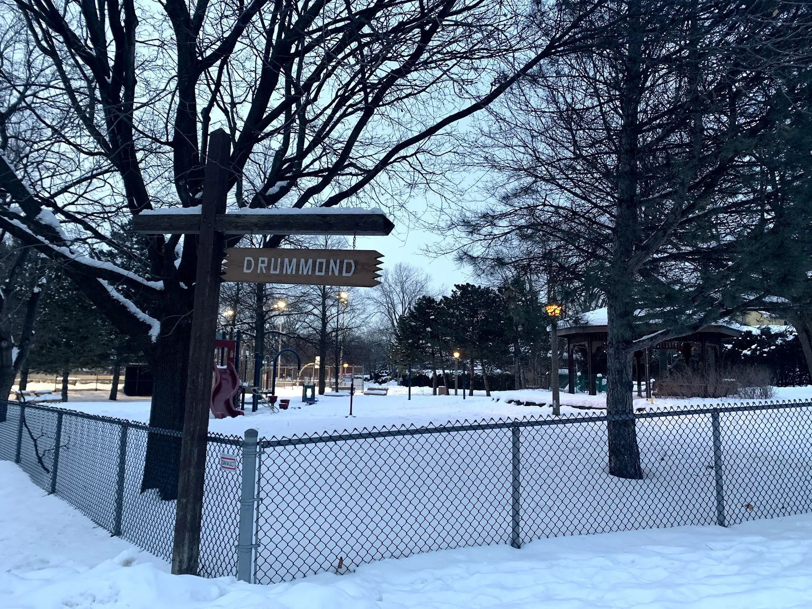 The Drummond Park sign and the fenced rink area under snow at dusk, with the playground and gazebo behind — Patinoire du parc Drummond (Beaconsfield)