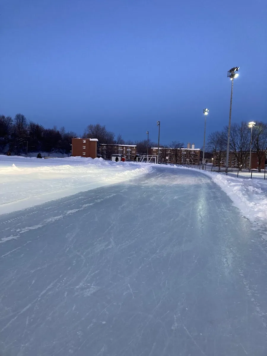L'anneau de glace entretenu du parc Ducharme à Sainte-Thérèse au crépuscule, avec des projecteurs et des immeubles d'habitation au-delà des bancs de neige — Patinoire du parc Ducharme