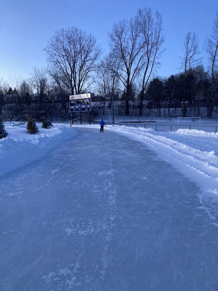 Un patineur seul au loin sur l'anneau de glace du parc Ducharme à Sainte-Thérèse au crépuscule, avec le tableau indicateur et des bancs de neige à côté de la glace — Patinoire du parc Ducharme