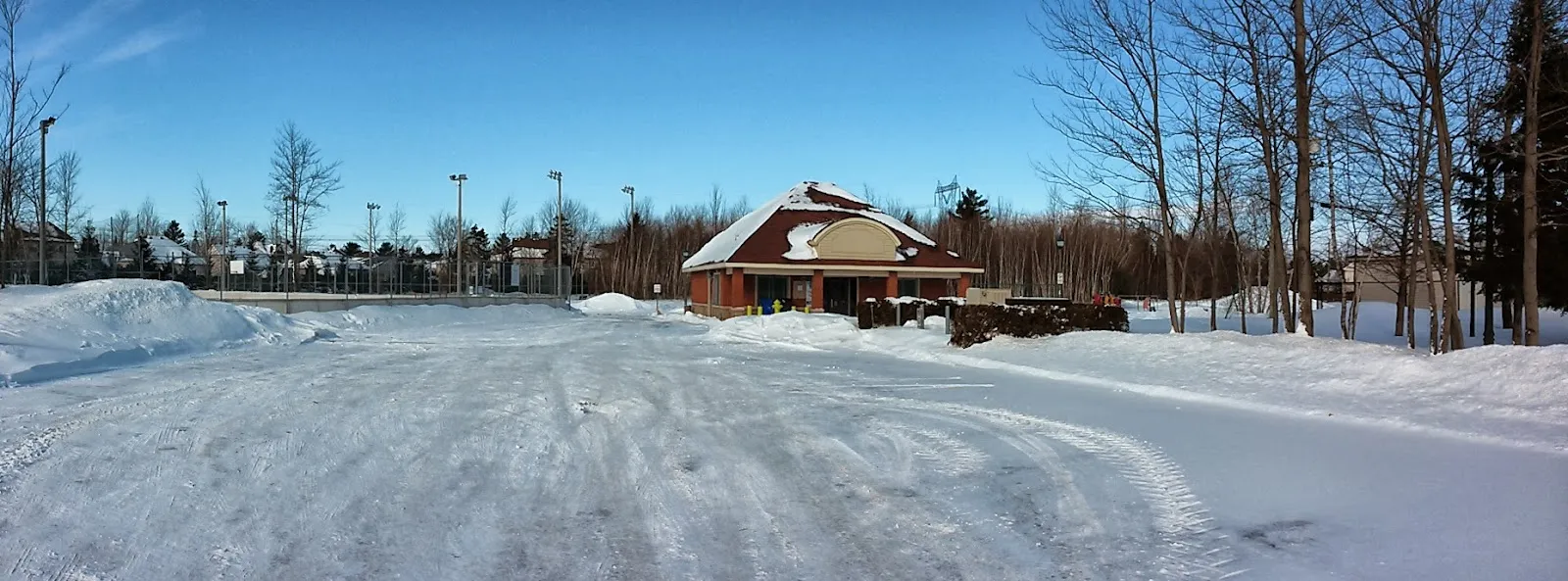 The park chalet at Parc Jacques-Beauchamp in Mirabel in winter, with the floodlit fenced rink area at the left under snow — Patinoire du parc Jacques-Beauchamp