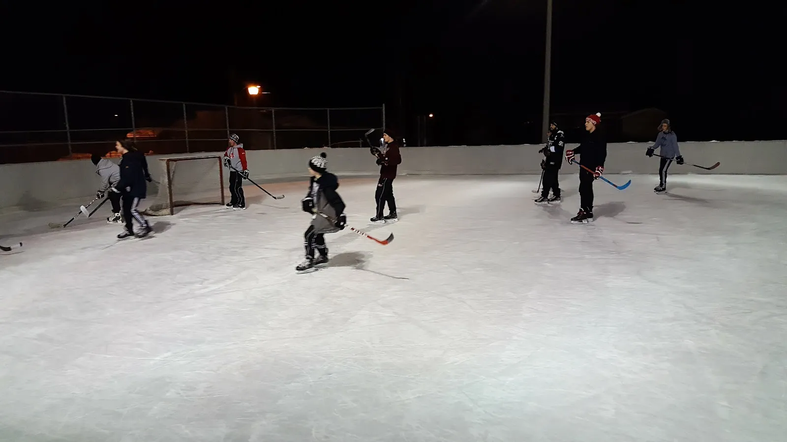A pick-up hockey game under floodlights on the boarded outdoor rink at Parc Lacroix in Mirabel at night, players at a distance — Patinoire du parc Lacroix