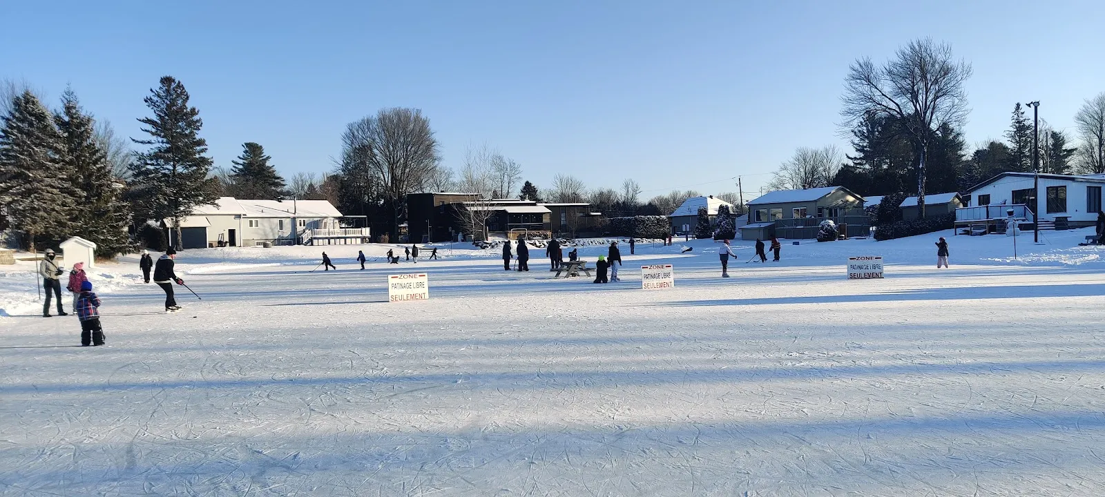 The lake rink at Parc Meilleur in Mirabel on a bright winter day, with skaters and hockey players at a distance and the lakeside houses beyond — Patinoire du parc Lucien-Meilleur