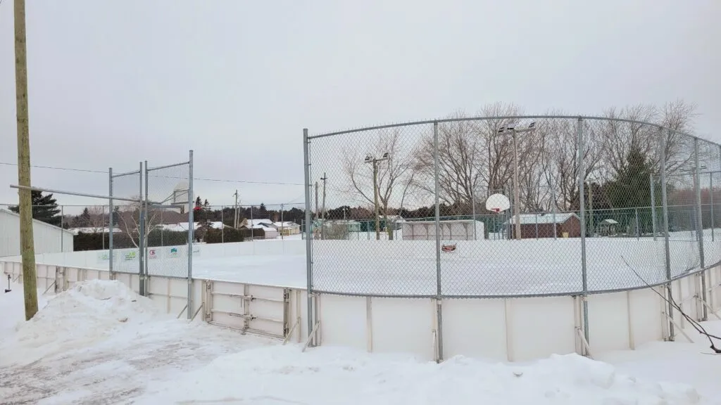The snow-edged boarded rink in the Sainte-Barbe municipal park under a grey winter sky, with a chain-link fence around it and no one on the ice. — Patinoire du parc municipal (Sainte-Barbe)