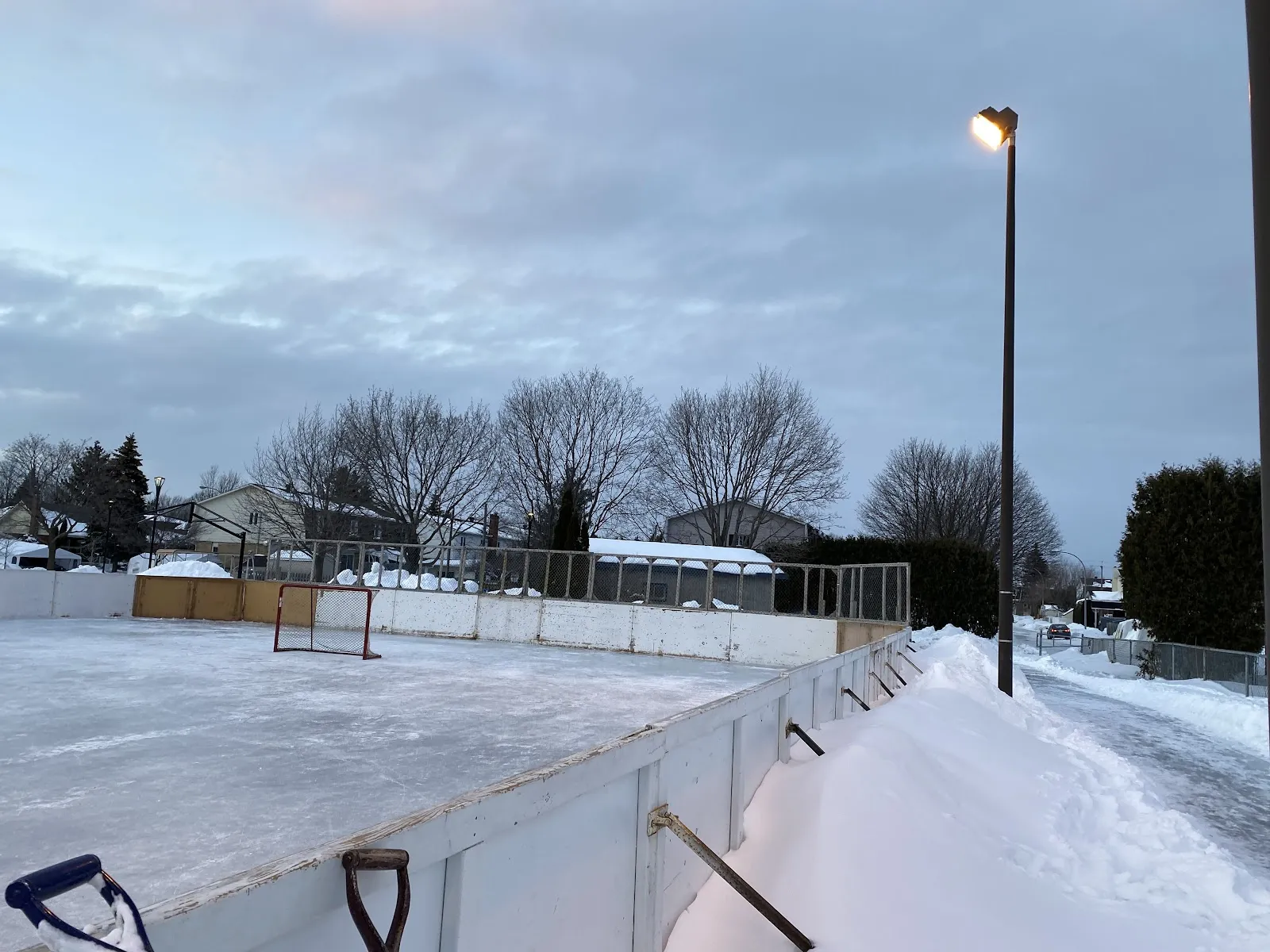 The boarded outdoor rink at Parc Olympia in Brossard at dusk, with goal, floodlight and neighbouring houses — Patinoire du parc Olympia