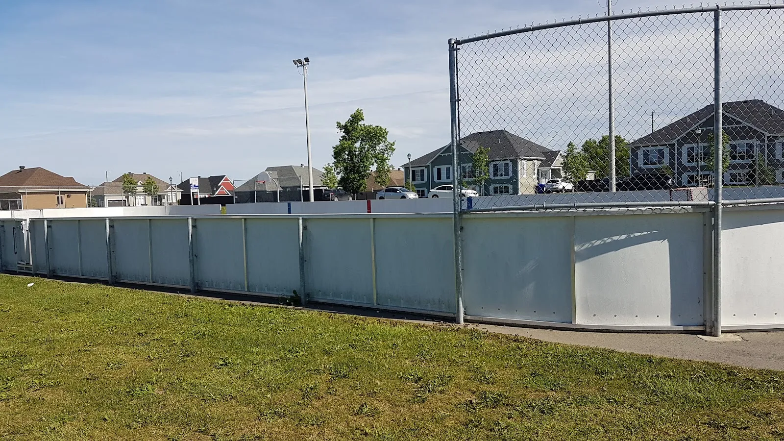 Les bandes blanches et la clôture de la patinoire extérieure du parc Raymond-Fortier à Mirabel vues depuis la pelouse en été, avec des maisons et un projecteur derrière, sans glace — Patinoire du parc Raymond-Fortier