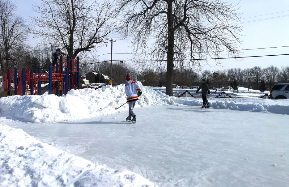 A lone skater on the cleared outdoor ice beside a playground, with snowbanks around the edge and bare trees behind. — Patinoire du parc Saint-Pierre (Pointe-des-Cascades)